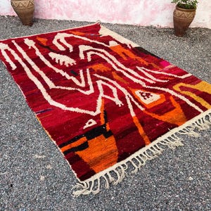 May include: A rectangular, hand-woven rug with a vibrant, abstract design. The rug features a rich palette of red, orange, and white, with fringe along the edges. The design includes geometric shapes and lines.