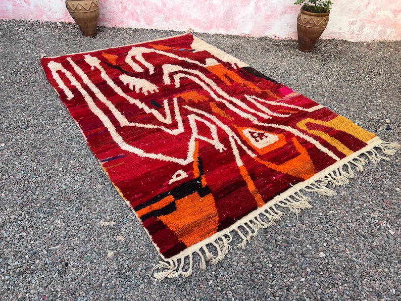May include: A rectangular, hand-woven rug with a vibrant, abstract design. The rug features a rich palette of red, orange, and white, with fringe along the edges. The design includes geometric shapes and lines.