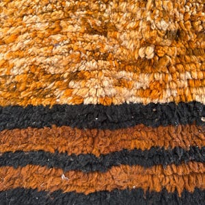 May include: Close-up of a textured rug with a mix of colors. The top section features a blend of orange and cream tones, transitioning to black and brown stripes. The bottom edge has a cream-colored fringe.