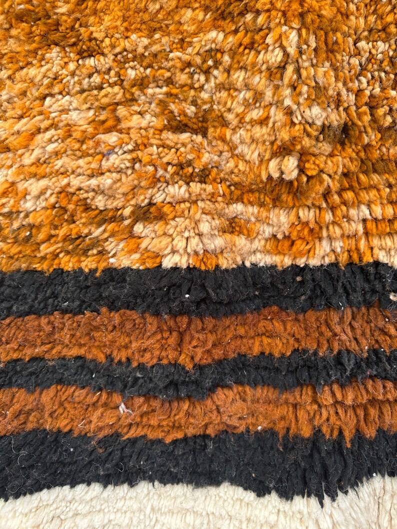 May include: Close-up of a textured rug with a mix of colors. The top section features a blend of orange and cream tones, transitioning to black and brown stripes. The bottom edge has a cream-colored fringe.
