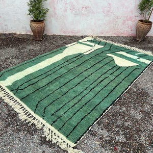 May include: A rectangular green rug with a cream-colored design and black stripes. The rug has a fringe on the edges. The rug is placed on a gravel surface. Two potted plants are in the background.