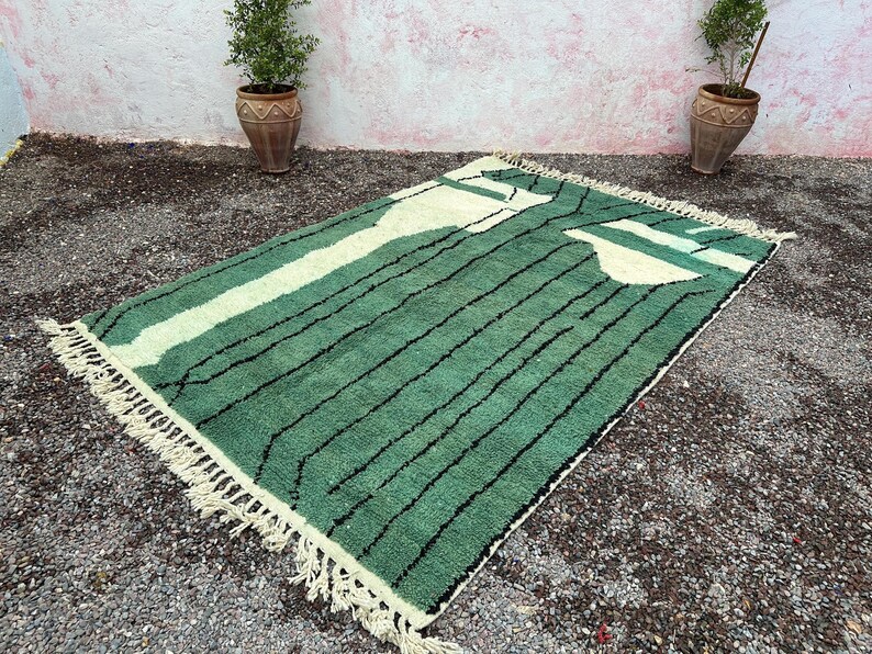 May include: A rectangular green rug with a cream-colored design and black stripes. The rug has a fringe on the edges. The rug is placed on a gravel surface. Two potted plants are in the background.