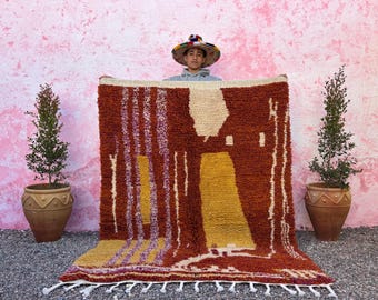 May include: A large, handwoven rug with a geometric pattern in shades of brown, yellow, and white. The rug has a fringe border and is being held up by a person wearing a colorful hat.