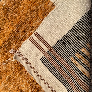 May include: Close-up of a textured, geometric rug with a mix of colors. The rug features a section of orange-brown, a cream-colored section, and a section with black and brown stripes. The rug's edge is visible, showing the woven construction.