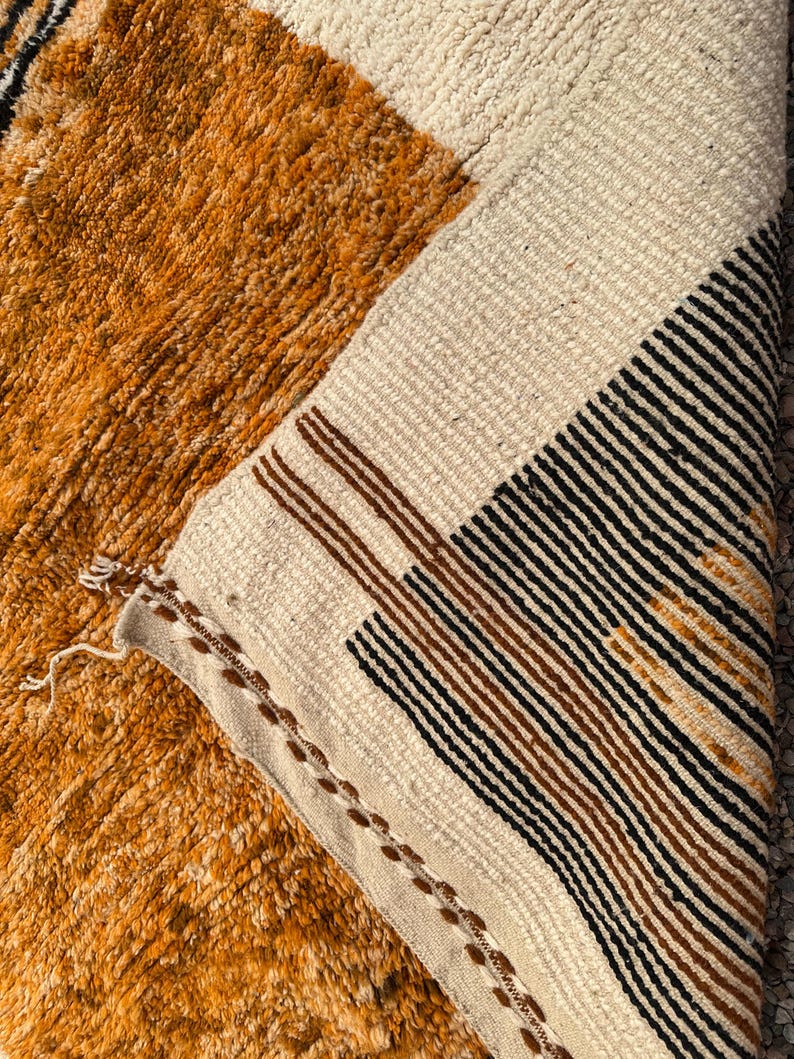 May include: Close-up of a textured, geometric rug with a mix of colors. The rug features a section of orange-brown, a cream-colored section, and a section with black and brown stripes. The rug's edge is visible, showing the woven construction.