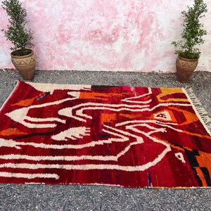 May include: A rectangular, hand-woven rug with a vibrant abstract design. The rug features a deep red background with white, orange, and black geometric patterns. The rug is displayed on a gray surface, with two potted plants in the background.