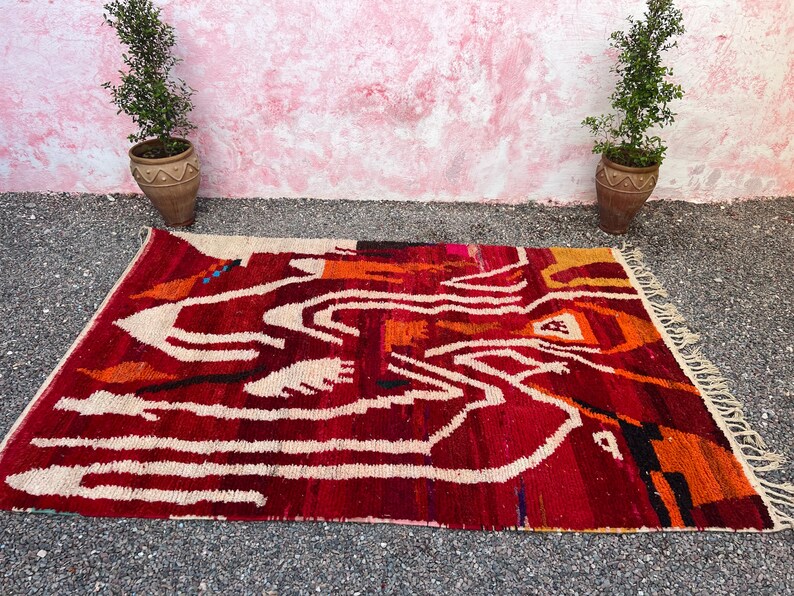 May include: A rectangular, hand-woven rug with a vibrant abstract design. The rug features a deep red background with white, orange, and black geometric patterns. The rug is displayed on a gray surface, with two potted plants in the background.