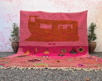 May include: A vibrant pink rug with an orange geometric design. The rug features a fringe along the bottom edge and is displayed against a textured pink wall. Two potted plants flank the rug.