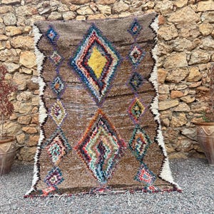 May include: A rectangular rug featuring a brown base with a large, central yellow diamond shape surrounded by smaller, multicolored geometric diamond patterns in shades of red, blue, green, and purple, finished with white fringed edges.