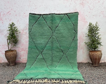 May include: A green rug with a diamond pattern and black outlines. The rug has a fringe at the bottom and is displayed against a pink and white wall. Two potted plants flank the rug.