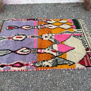 May include: A rectangular, hand-woven rug with a vibrant, geometric design. The rug features a color palette of purple, orange, yellow, pink, and cream, with black and white accents. The rug has a fringed edge and is likely made of wool.