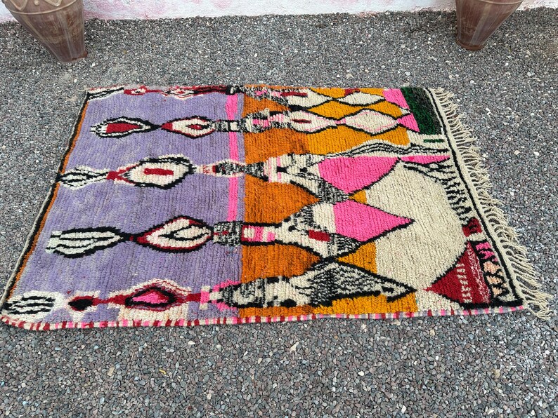 May include: A rectangular, hand-woven rug with a vibrant, geometric design. The rug features a color palette of purple, orange, yellow, pink, and cream, with black and white accents. The rug has a fringed edge and is likely made of wool.
