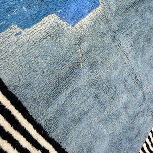 May include: A close-up of a textured rug with a geometric design. The rug features shades of blue and gray, with a section of black and white stripes along the edge. The rug's texture is visible, suggesting a soft, woven material.