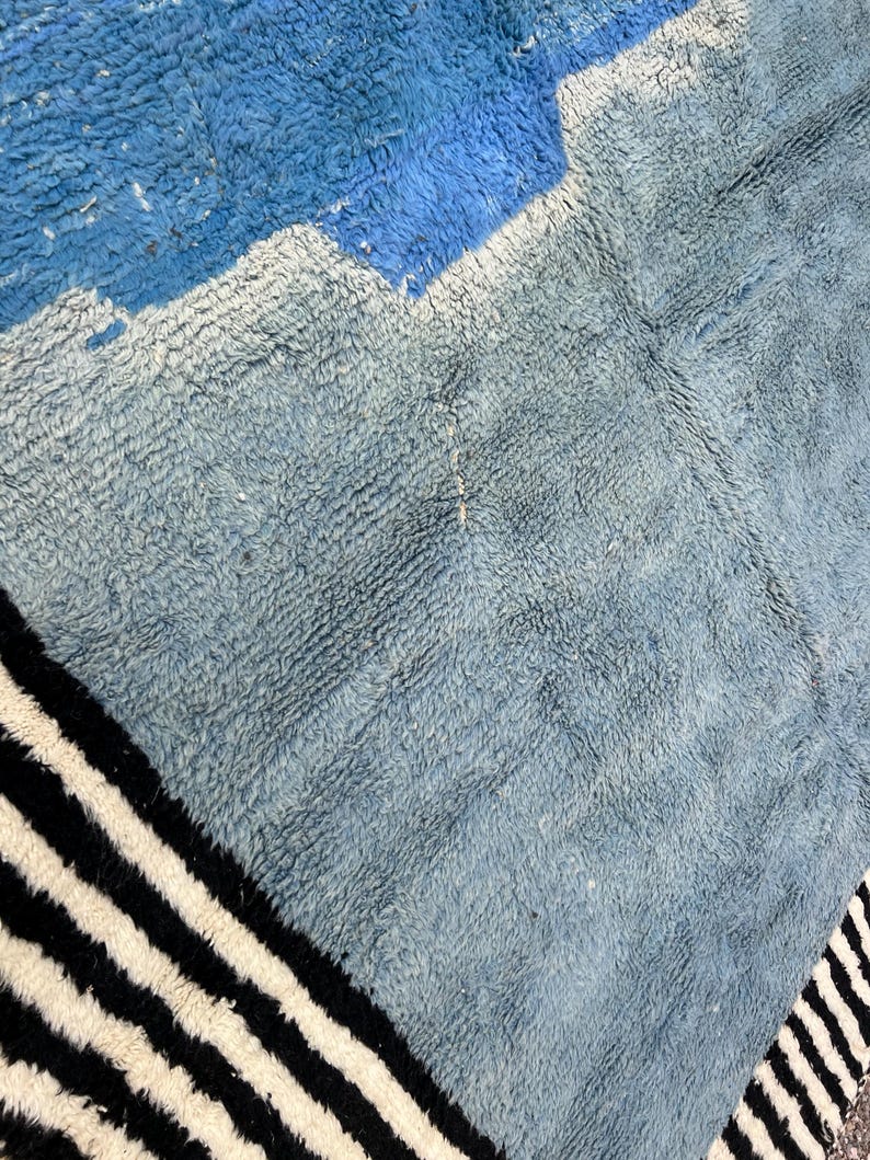 May include: A close-up of a textured rug with a geometric design. The rug features shades of blue and gray, with a section of black and white stripes along the edge. The rug's texture is visible, suggesting a soft, woven material.