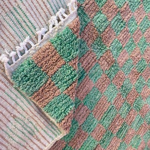 May include: A close-up of a green and brown checkered bath mat with a white fringe border. The mat is made of a soft, fluffy material.