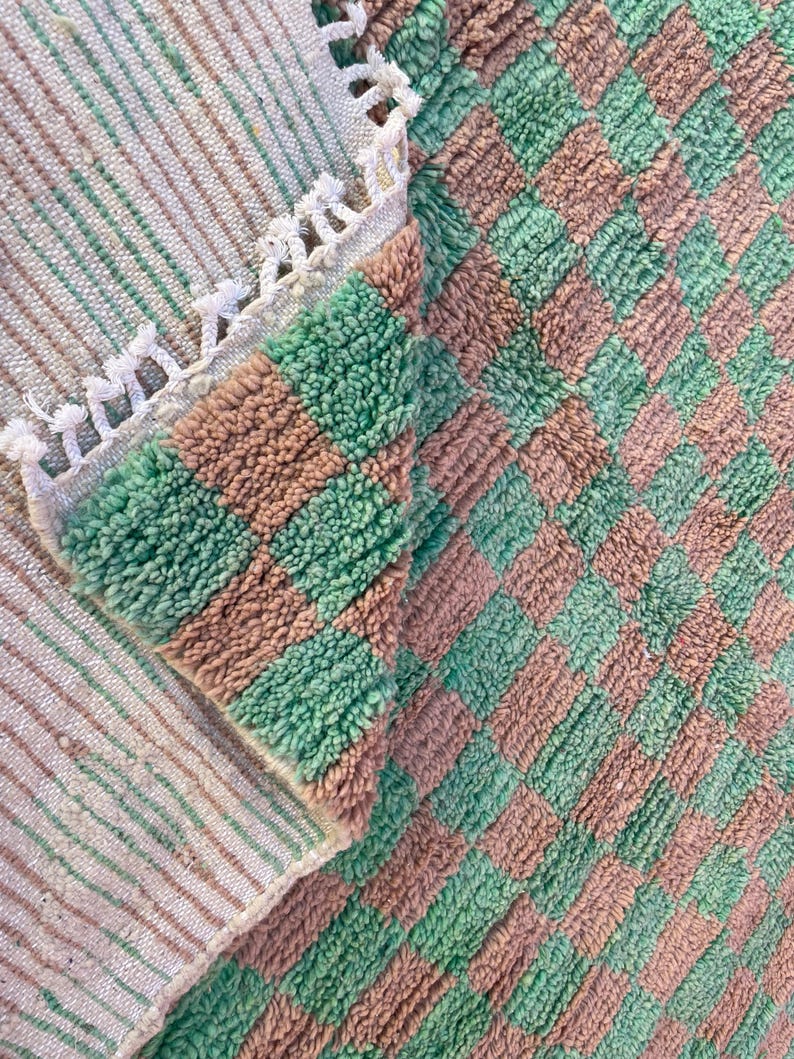 May include: A close-up of a green and brown checkered bath mat with a white fringe border. The mat is made of a soft, fluffy material.