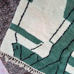 May include: A close-up of a section of a rug with a geometric design. The rug features a cream-colored background with green and black geometric shapes. The rug has a fringe along one edge.