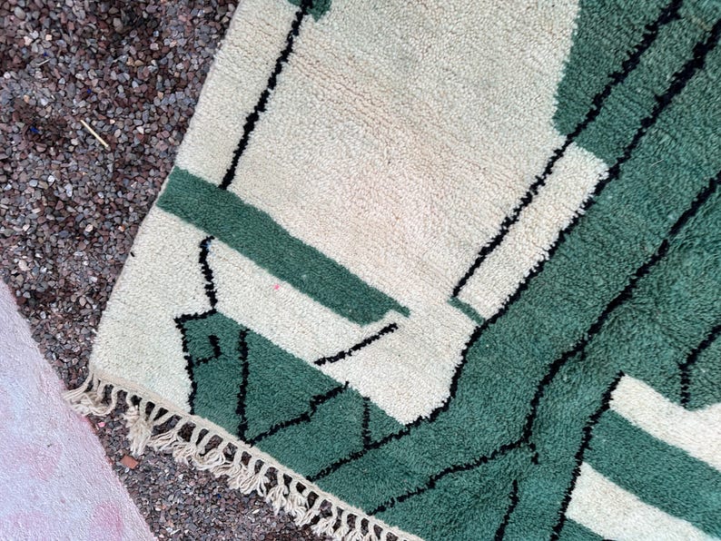 May include: A close-up of a section of a rug with a geometric design. The rug features a cream-colored background with green and black geometric shapes. The rug has a fringe along one edge.