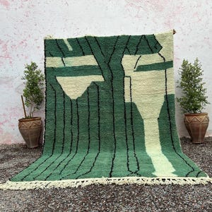 May include: A green and cream-colored rug with a geometric design. The rug features a pattern of vertical black lines and abstract shapes in cream and green. The rug has a fringe along the bottom edge.