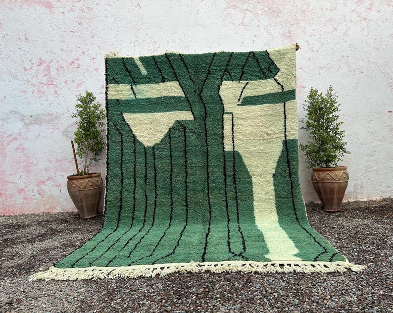 May include: A green and cream-colored rug with a geometric design. The rug features a pattern of vertical black lines and abstract shapes in cream and green. The rug has a fringe along the bottom edge.