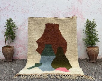 May include: A beige and brown woven rug with an abstract design featuring shapes in brown, green, blue, and pink. The rug has a fringed edge and is displayed outdoors.