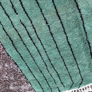 May include: A section of a green rug with black vertical stripes. The rug has a textured surface and a white fringe along the bottom edge. A section of small, brown pebbles is visible on the left side.
