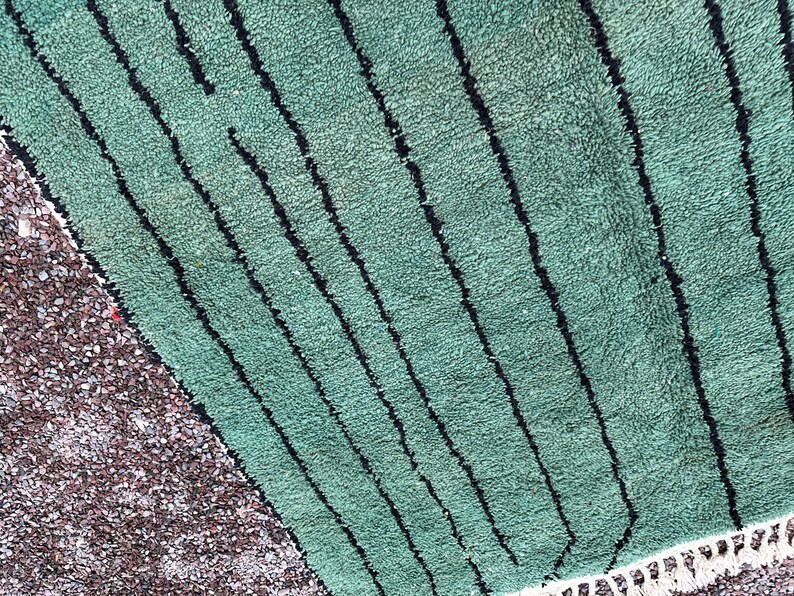 May include: A section of a green rug with black vertical stripes. The rug has a textured surface and a white fringe along the bottom edge. A section of small, brown pebbles is visible on the left side.