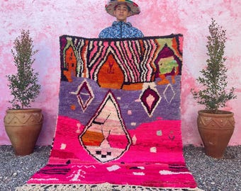 May include: A colorful Moroccan rug with geometric patterns in shades of pink, purple, orange, and black. The rug has a fringe border and is being held up by a person wearing a hat.