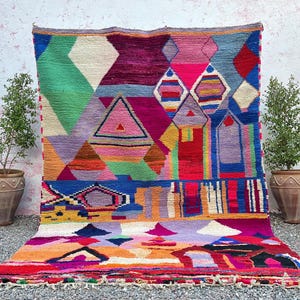 Può includere: Un vivace tappeto geometrico con un design colorato. Il tappeto presenta un mix di forme, tra cui diamanti, triangoli e rettangoli, nelle tonalità di verde, rosa, viola e arancione. Il tappeto è esposto su una superficie di ghiaia con piante in vaso su entrambi i lati.