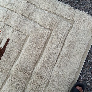 May include: Cream-colored, textured rug with a geometric design. The rug features a series of rectangular shapes and borders, with accents of brown, orange, and black. The rug's texture appears soft and plush, suitable for a living space.