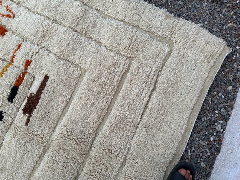 May include: Cream-colored, textured rug with a geometric design. The rug features a series of rectangular shapes and borders, with accents of brown, orange, and black. The rug's texture appears soft and plush, suitable for a living space.