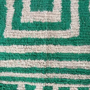 Pode incluir: Close-up de um tapete com padr&atilde;o geom&eacute;trico verde e branco sujo. O tapete apresenta um design repetitivo de quadrados e listras horizontais. A textura parece macia e felpuda.