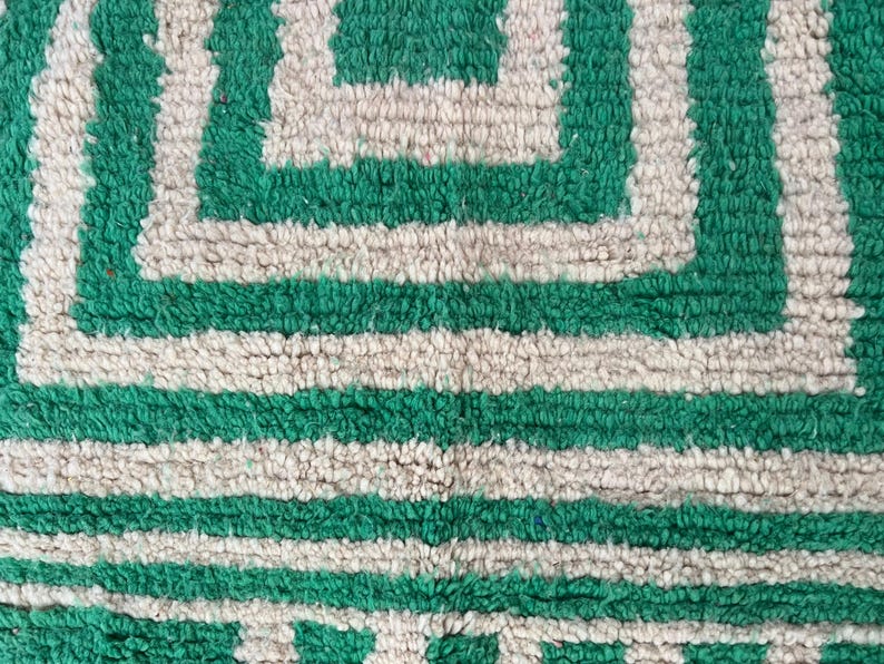 Pode incluir: Close-up de um tapete com padr&atilde;o geom&eacute;trico verde e branco sujo. O tapete apresenta um design repetitivo de quadrados e listras horizontais. A textura parece macia e felpuda.