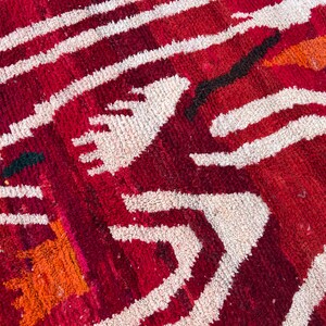 May include: Close-up of a red and white patterned rug. The rug features a geometric design with thick white lines and shapes against a deep red background. Orange and black accents add contrast to the design. The rug has a textured appearance.