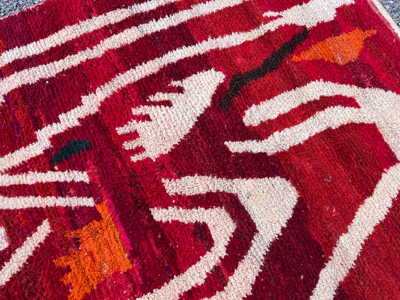 May include: Close-up of a red and white patterned rug. The rug features a geometric design with thick white lines and shapes against a deep red background. Orange and black accents add contrast to the design. The rug has a textured appearance.