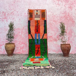 May include: A long, narrow rug with a geometric pattern in shades of green, orange, red, blue, and black. The rug has a fringe on the bottom.