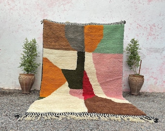 May include: A colorful, geometric rug with a textured surface. The rug features a patchwork design with shades of brown, orange, green, pink, and cream. The rug has a black and white fringed edge. Two potted plants flank the rug.