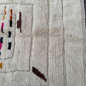May include: Close-up of a cream-colored, textured rug with a geometric design. The rug features vertical stripes and colorful abstract shapes in brown, orange, black, and pink. The rug's texture appears soft and plush.