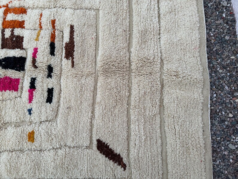 May include: Close-up of a cream-colored, textured rug with a geometric design. The rug features vertical stripes and colorful abstract shapes in brown, orange, black, and pink. The rug's texture appears soft and plush.