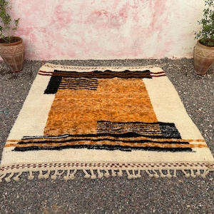 May include: A rectangular, hand-woven rug with a cream-colored background and geometric patterns in shades of brown, black, and orange. The rug features a fringe along the bottom edge and is suitable for home decor.