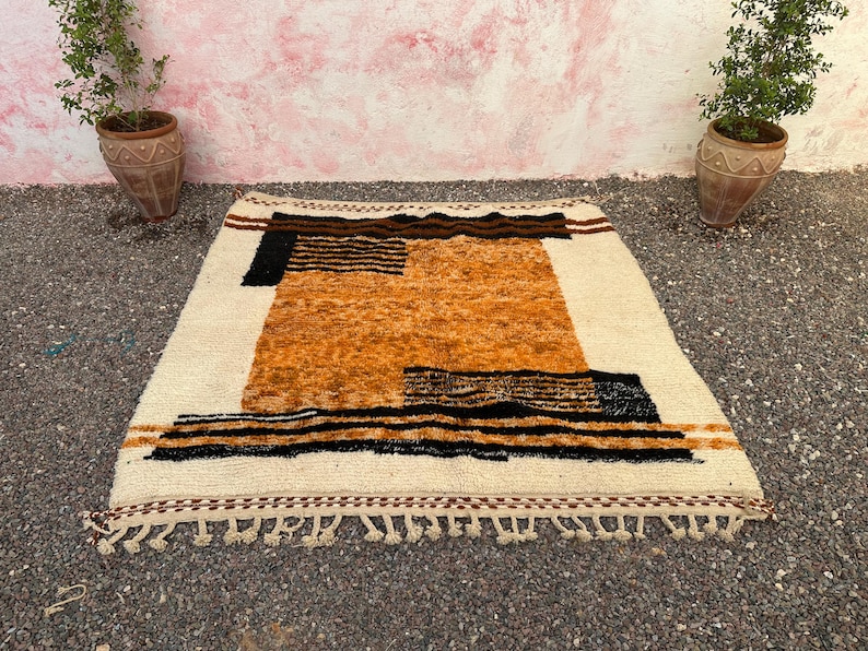 May include: A rectangular, hand-woven rug with a cream-colored background and geometric patterns in shades of brown, black, and orange. The rug features a fringe along the bottom edge and is suitable for home decor.