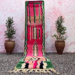 May include: A colourful runner rug with a pink central design, bordered by green and black zigzag patterns. The rug features a geometric pattern with cream and black accents, and a fringe at the bottom. The rug is displayed on a bed of gravel.