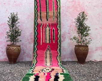 May include: A vibrant, rectangular runner rug with a pink central design, bordered by green and black zigzag patterns. The rug features a geometric pattern with cream and black accents, and a fringe at the bottom. The rug is displayed on a bed of gravel.