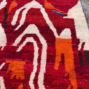 May include: Close-up of a vibrant, hand-woven rug with a rich red background. The rug features bold, abstract designs in white, orange, and brown. The texture is thick and plush, suggesting a high-quality, durable construction. The rug's design is reminiscent of tribal art.