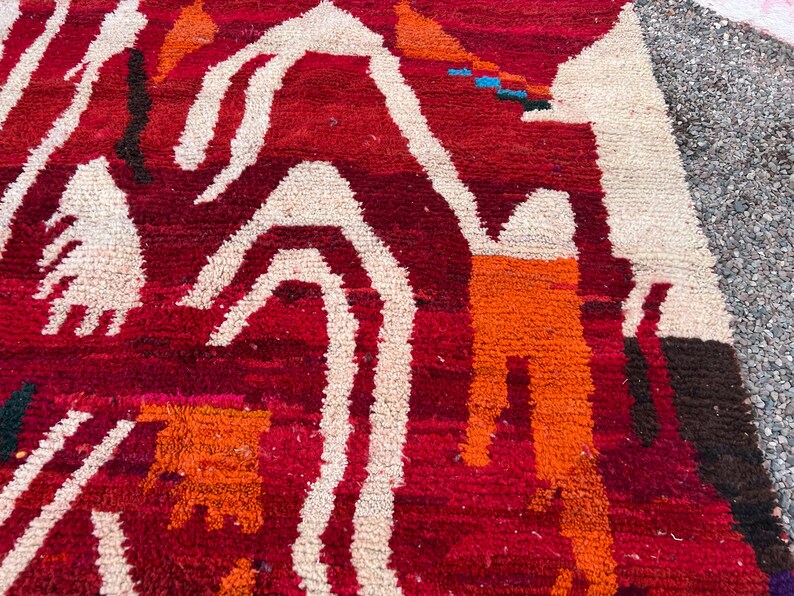 May include: Close-up of a vibrant, hand-woven rug with a rich red background. The rug features bold, abstract designs in white, orange, and brown. The texture is thick and plush, suggesting a high-quality, durable construction. The rug's design is reminiscent of tribal art.