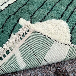May include: A close-up of a green and cream-colored rug with a geometric design. The rug features a textured, plush pile with black lines and a fringed edge. The rug is folded back, revealing the striped underside.