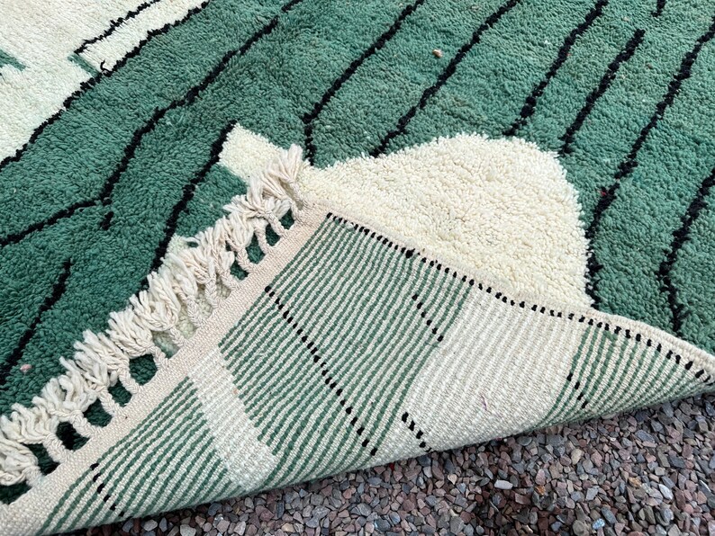 May include: A close-up of a green and cream-colored rug with a geometric design. The rug features a textured, plush pile with black lines and a fringed edge. The rug is folded back, revealing the striped underside.