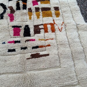 May include: Cream-colored, textured rug with a geometric design. The rug features a repeating pattern of rectangles and squares in shades of black, brown, pink, orange, and yellow. The rug's texture is thick and plush, with a soft, inviting appearance.