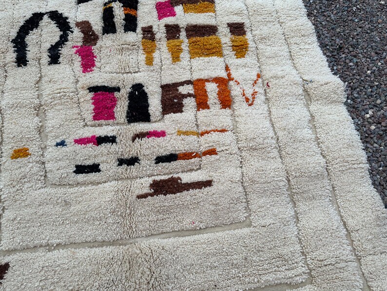 May include: Cream-colored, textured rug with a geometric design. The rug features a repeating pattern of rectangles and squares in shades of black, brown, pink, orange, and yellow. The rug's texture is thick and plush, with a soft, inviting appearance.