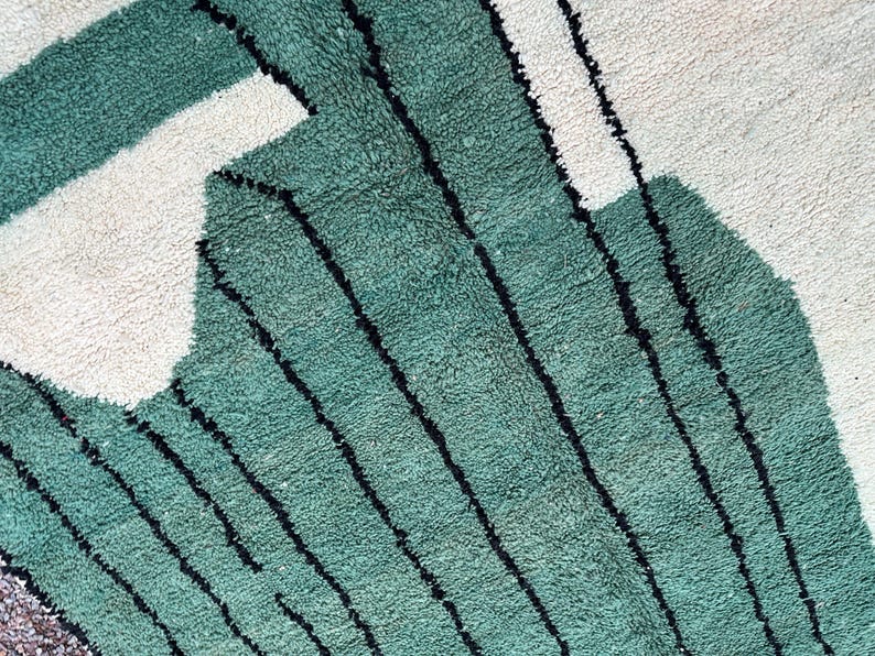 May include: Close-up of a textured rug with a geometric design. The rug features a teal green color with black stripes and white shapes. The rug has a soft, plush appearance.